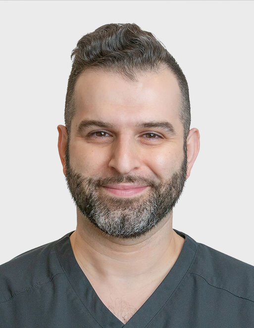 A man with short, dark hair and a trimmed beard, wearing a dark scrub top, smiles at the camera against a plain white background.