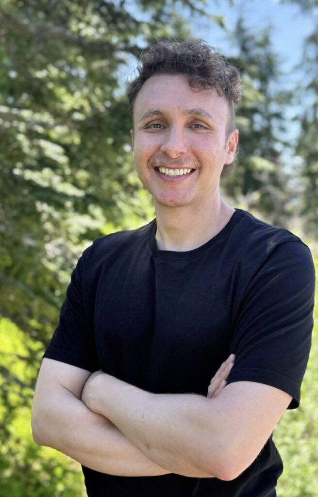 A person with short curly hair, wearing a black t-shirt, stands outdoors in North Vancouver Lonsdale with arms crossed and smiles at the camera. Green trees and sunlight are in the background.