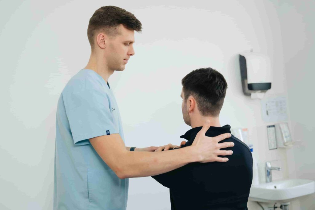A healthcare professional in scrubs examines a seated patient's shoulders in a medical office, recommending top shoulder pain physio exercises Coquitlam patients trust.
