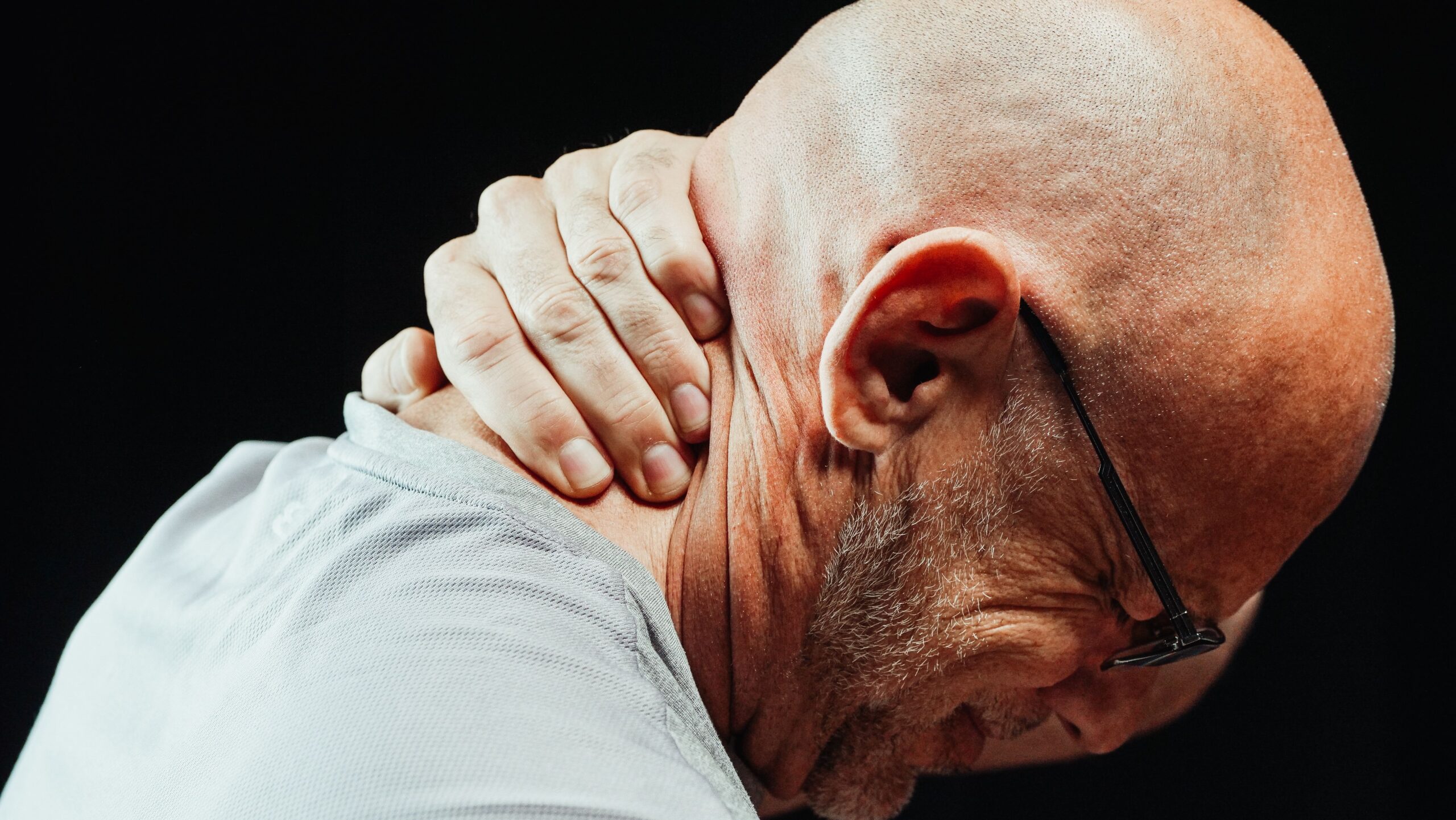 A bald man wearing glasses holds the back of his neck, appearing to experience chronic pain, possibly seeking relief through holistic physiotherapy.