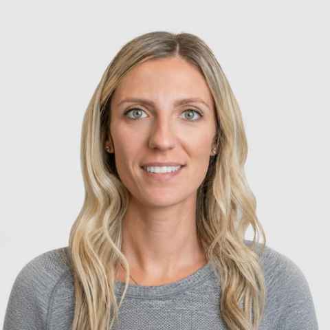 Woman with long blonde hair and gray sweater, facing forward and smiling slightly, against a plain light gray background.