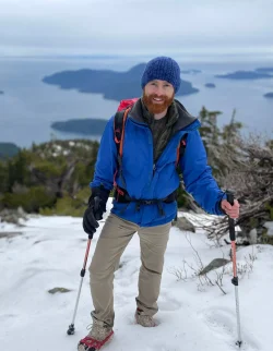 A person in a blue jacket and knit hat stands on a snowy mountain slope with hiking poles, overlooking a scenic view of water and islands, reminiscent of an adventure by Thomas Scarr.