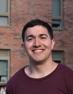 Thomas Scarr, a young man with short dark hair, wearing a maroon shirt, smiles in front of a brick building with several windows.