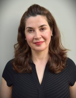 Joan Eustaquio, with wavy brown hair, wears a black short-sleeved top as she stands before a plain light-colored background, gazing at the camera with a neutral expression.