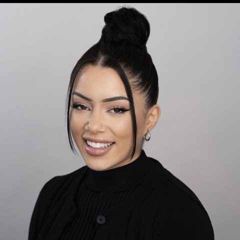 With long black hair in a bun and hoop earrings paired with a black sweater, she exudes confidence and warmth. Her smile suggests a passion for Active Rehab, radiating positivity against the gray background.