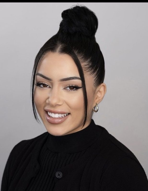 With long black hair in a bun and hoop earrings paired with a black sweater, she exudes confidence and warmth. Her smile suggests a passion for Active Rehab, radiating positivity against the gray background.