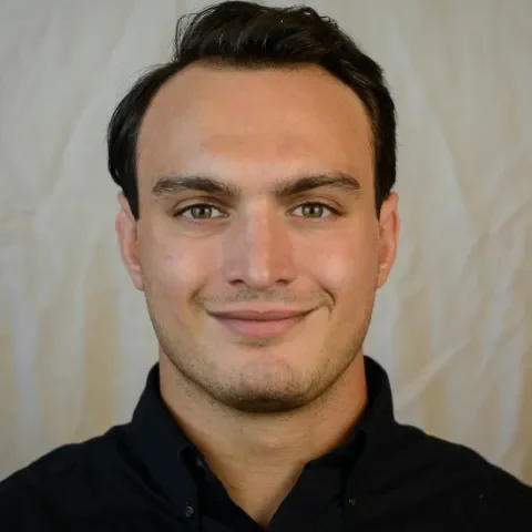 A man with short dark hair and light skin is facing the camera, wearing a black collared shirt. The plain, light-colored background gives a professional look, suitable for a Mobile Physiotherapy profile photo.
