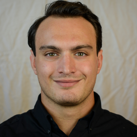 A young man with short dark hair and a slight smile, wearing a black button-up shirt, stands against a plain, off-white background, embodying the approachable and professional demeanor of mobile physiotherapy.